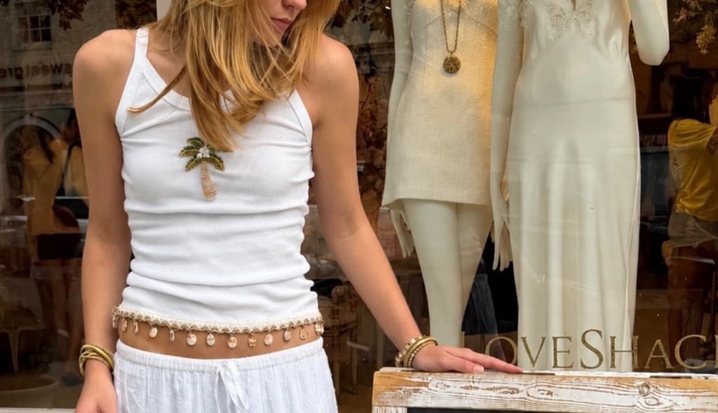 Coast Collection look: white tank with a palm-tree patch, shell belt, and cream trousers in front of a boutique window